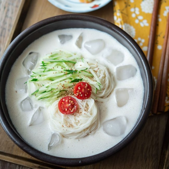 Healthy Korean kongguksu cold soybean noodle soup topped with cucumber and tomato, served with ice cubes for a refreshing summer dish.