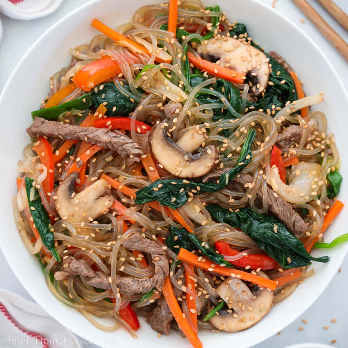 Korean Japchae glass noodles stir-fried with beef, spinach, carrots, bell peppers, and shiitake mushrooms topped with sesame seeds in a white bowl.